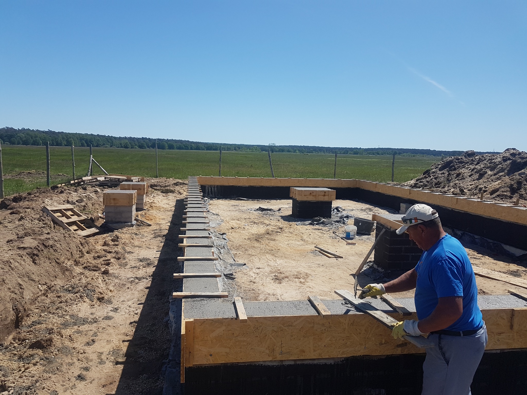 Pracownik w niebieskiej koszulce i czapce z daszkiem wyrównuje beton w szalunku fundamentu domu jednorodzinnego na tle zielonego pola i błękitnego nieba.