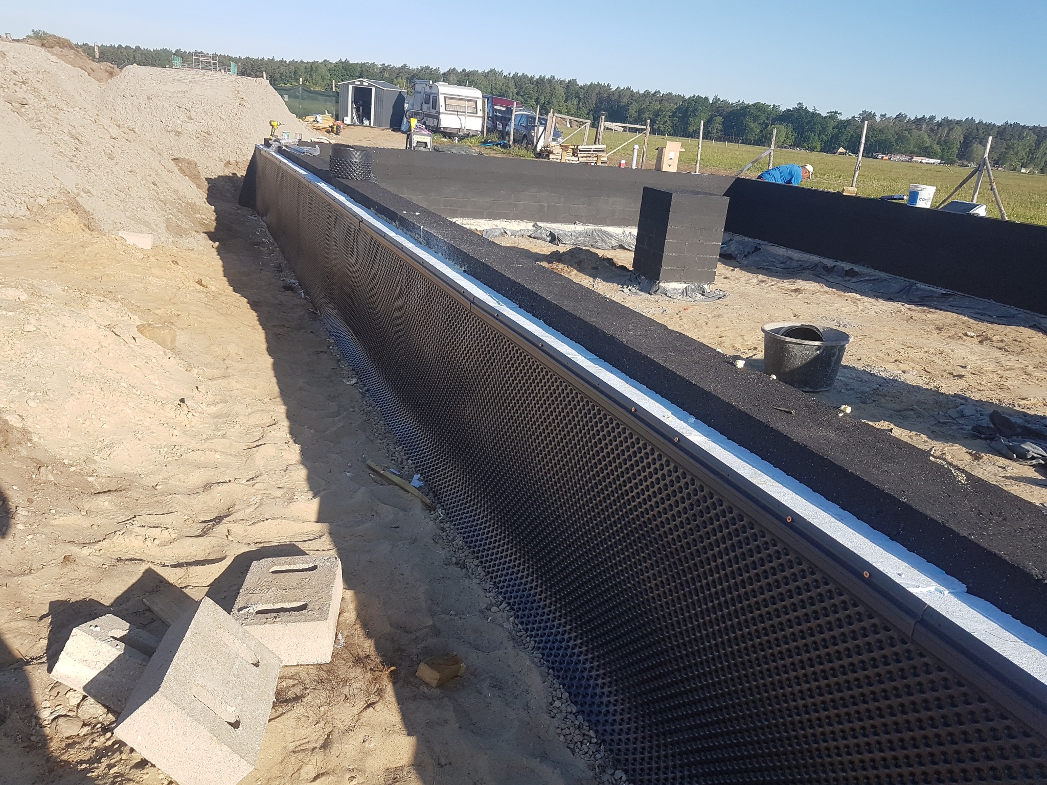 Przygotowanie fundamentów pod budowę domu: zabezpieczona ściana fundamentowa z izolacją pionową, folią kubełkową i warstwą ocieplenia, widoczne bloczki fundamentowe i zasypana ziemia