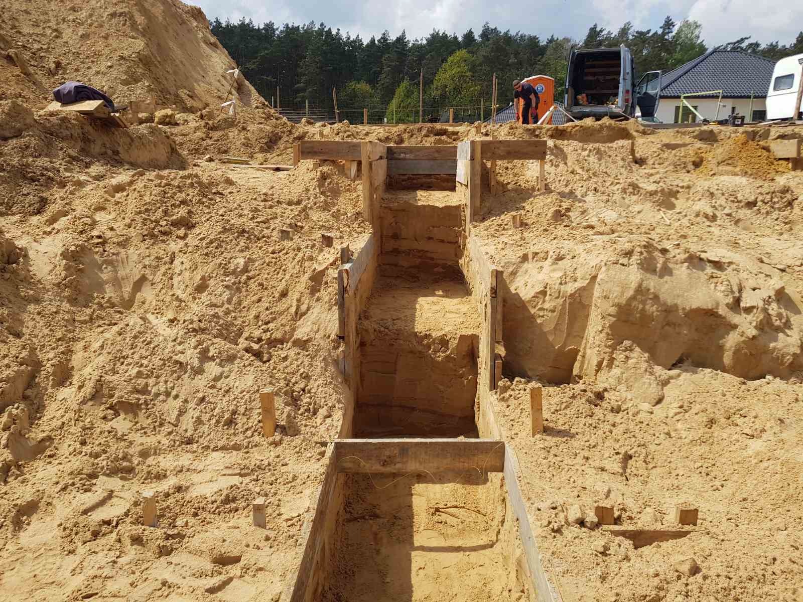Wykop pod fundamenty z deskowaniem, widoczny pracownik w tle i zaparkowany bus.