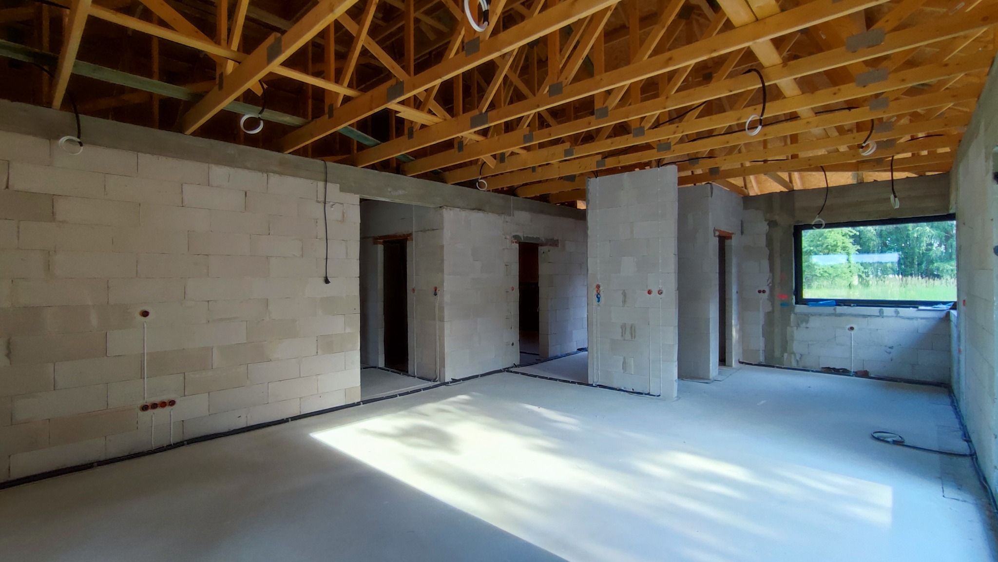 Surowy stan wnętrza domu w trakcie budowy, widoczne ściany z bloczków, drewniana konstrukcja dachu, okablowanie elektryczne w peszlach na ścianach i pod sufitem, oraz puszki elektryczne podtynkowye.