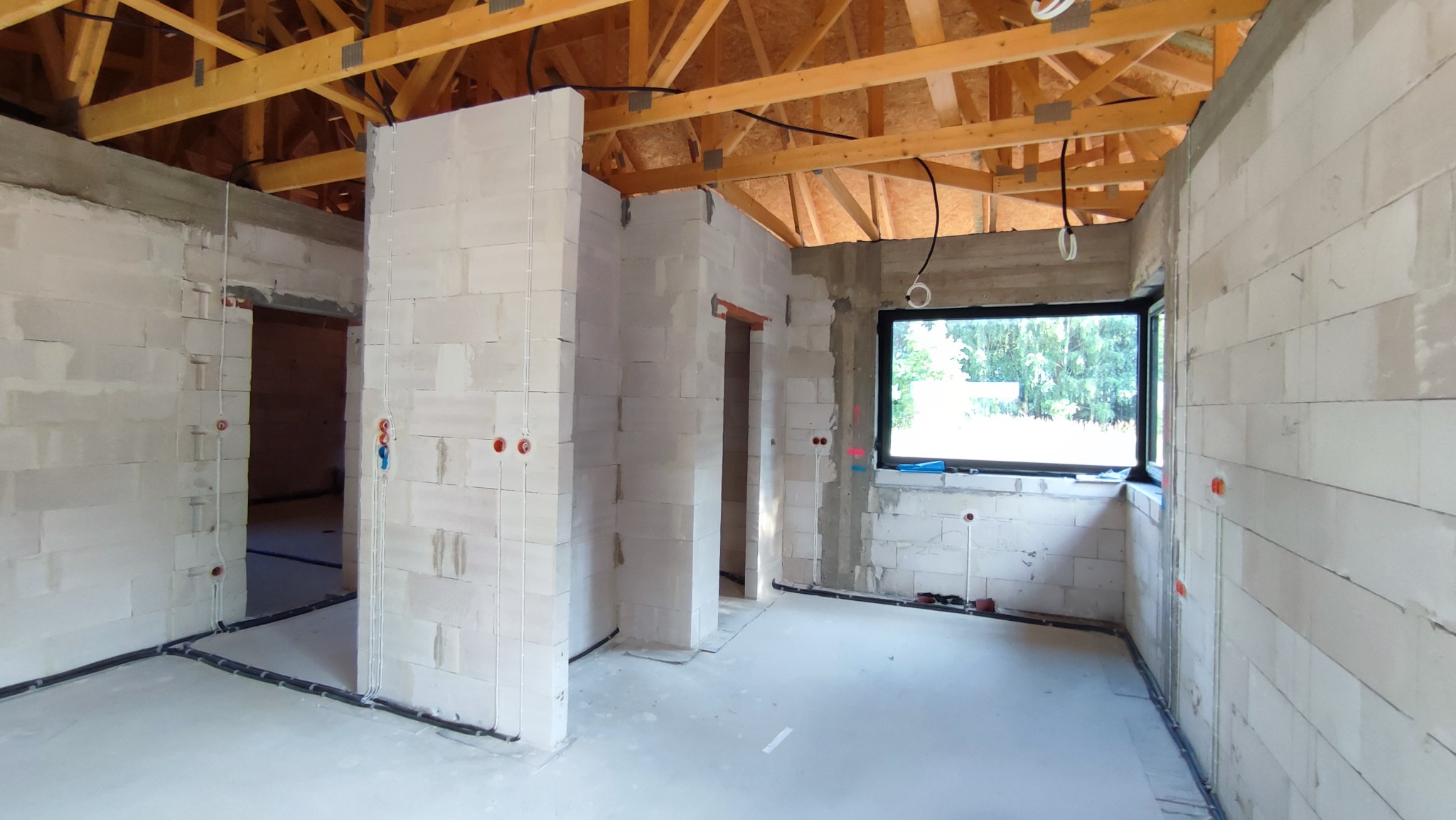 Wnętrze domu w trakcie budowy z widocznymi przewodami elektrycznymi w ścianach z betonu komórkowego oraz rurami instalacyjnymi przy podłodze, drewniana konstrukcja dachu.