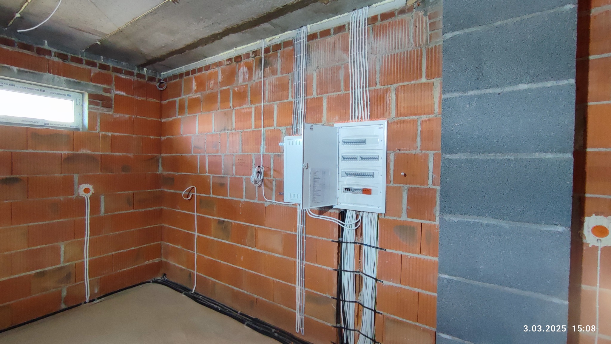 Rozdzielnica elektryczna z otwartymi drzwiczkami na ścianie z cegły dziurawki, widoczne kable w peszlach i podłączenia w ścianie, surowy stan wykończenia wnętrza.