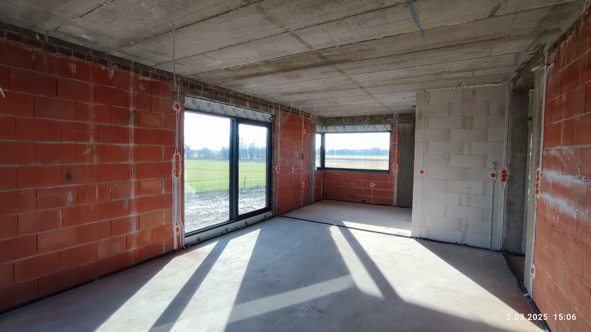 Surowy stan wnętrza budynku z widoczną instalacją elektryczną w bruzdach ścian z czerwonej cegły i betonu, okna z widokiem na zielone pole.