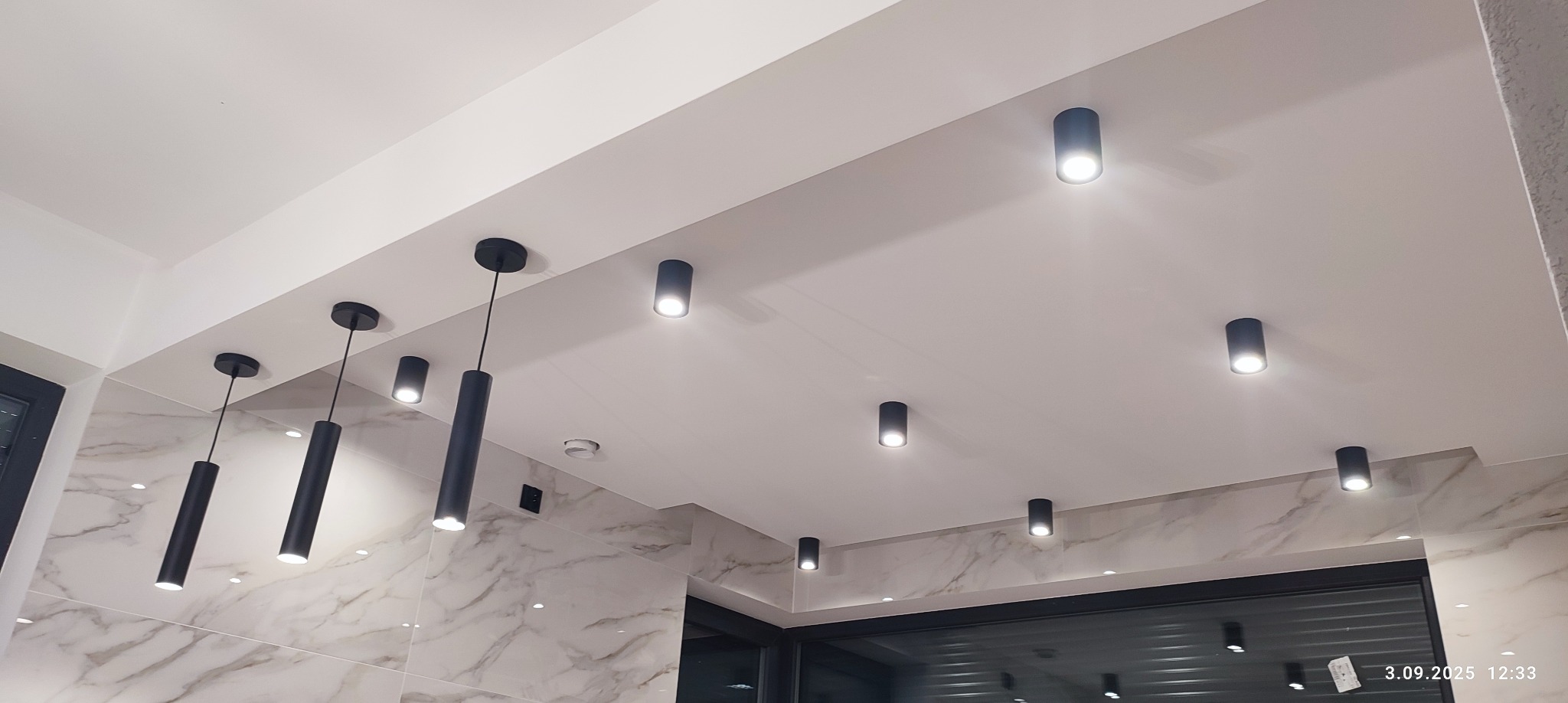 Nowoczesne oświetlenie sufitowe z czarnymi, cylindrycznymi lampami wiszącymi i punktowymi w białym wnętrzu z marmurowym wykończeniem ścian.
