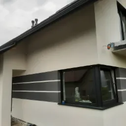 Fragment elewacji domu po tynkowaniu i malowaniu, z szarymi pasami dekoracyjnymi, widoczne okno z czarną ramą, rynna i fragment dachu. Na ziemi leżą złożone rusztowania.