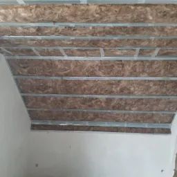 Skośny sufit poddasza z widoczną izolacją z wełny mineralnej pomiędzy metalowymi profilami konstrukcyjnymi, mocowanymi taśmą klejącą, w trakcie prac wykończeniowych.