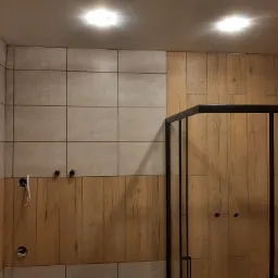 Ściana łazienki wyłożona częściowo szarymi płytkami, częściowo panelami imitującymi drewno, z widoczną czarną ramą kabiny prysznicowej i niedokończonymi instalacjami hydraulicznymi.