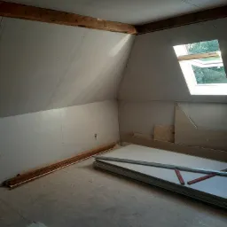 Poddasze z oknem dachowym, częściowo wykończone płytami kartonowo-gipsowymi, widoczne drewniane belki stropowe i przygotowane płyty oraz profile do dalszego montażu.