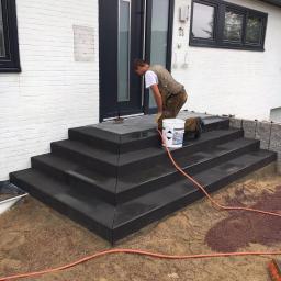 Truszkowski - Pracownik w stroju roboczym impregnuje nowo ułożone, ciemnoszare, modułowe schody przed wejściem do domu z białą cegłą, widoczny pomarańczowy kabel i wiadro z preparatem.