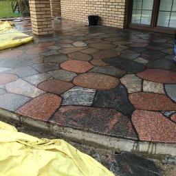 Truszkowski - Fragment tarasu z nieregularnych kamieni w odcieniach szarości i czerwieni, zabezpieczony żółtą folią budowlaną, widoczne narzędzia malarskie i wiadro, ceglane kolumny podtrzymujące zadaszenie.