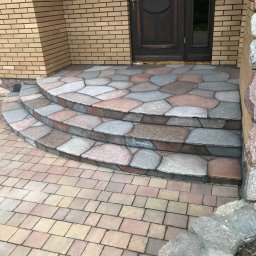 Truszkowski - Trzy stopnie wejściowe wyłożone nieregularnymi płytami kamiennymi w odcieniach szarości i różu, prowadzące do ciemnych drzwi wejściowych w budynku z cegły, z fragmentem chodnika z kostki brukowej...