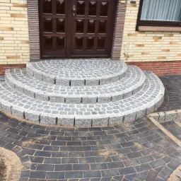 Półokrągłe schody wejściowe z kostki granitowej w odcieniach szarości, prowadzące do ciemnych, brukowanych ścieżek, z widocznymi drobnymi pozostałościami piasku po fugowaniu.