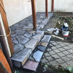 Truszkowski - Kamienny taras z nieregularnych płyt i schodami, widoczne drewniane podpory zadaszenia, obok rabata z roślinami i narzędzia budowlane.