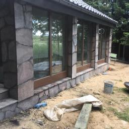 Truszkowski - Kamienna elewacja budynku z dużymi oknami i schodami wejściowymi, w trakcie prac wykończeniowych na zewnątrz widać narzędzia i materiały budowlane.