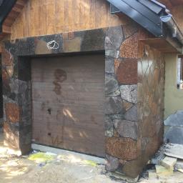 Truszkowski - Brama garażowa z drewnianym panelem, otoczona nieregularnym kamieniem elewacyjnym w odcieniach brązu i szarości, z fragmentem ściany obok okna w trakcie wykończenia.