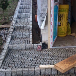 Truszkowski - Schody zewnętrzne z kostki granitowej prowadzące w górę, częściowo ułożone, z widocznymi elementami konstrukcyjnymi budynku w tle i roboczą rękawicą obok wejścia.