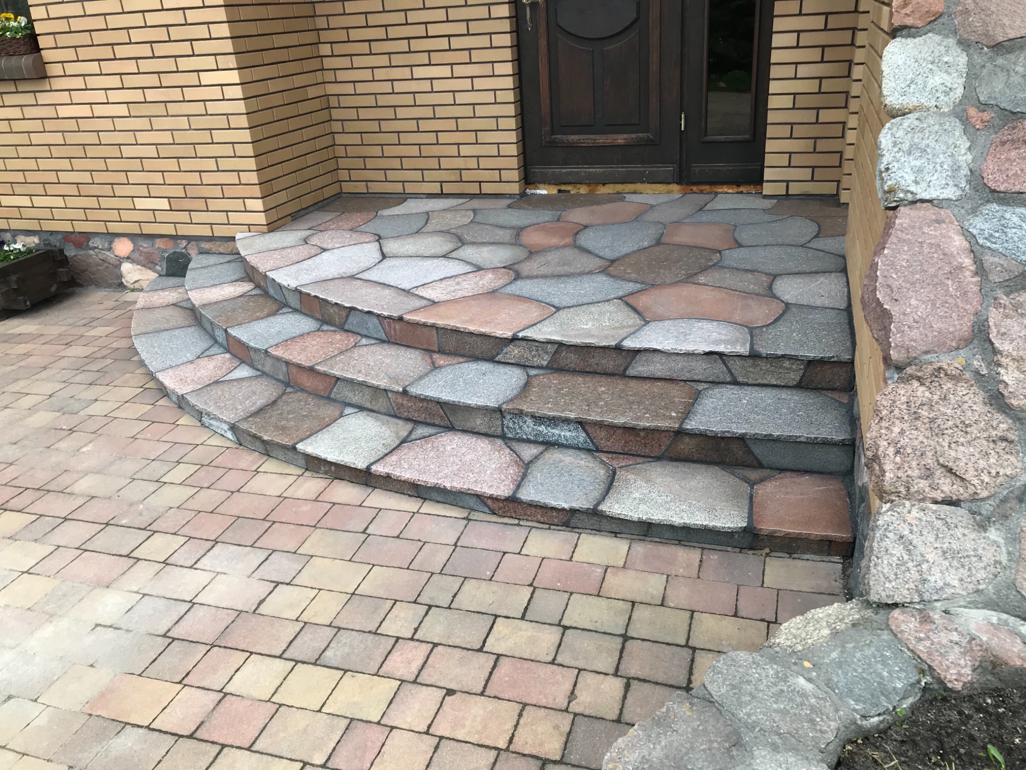 Trzy stopnie wejściowe wyłożone nieregularnymi płytami kamiennymi w odcieniach szarości i różu, prowadzące do ciemnych drzwi wejściowych w budynku z cegły, z fragmentem chodnika z kostki brukowej...
