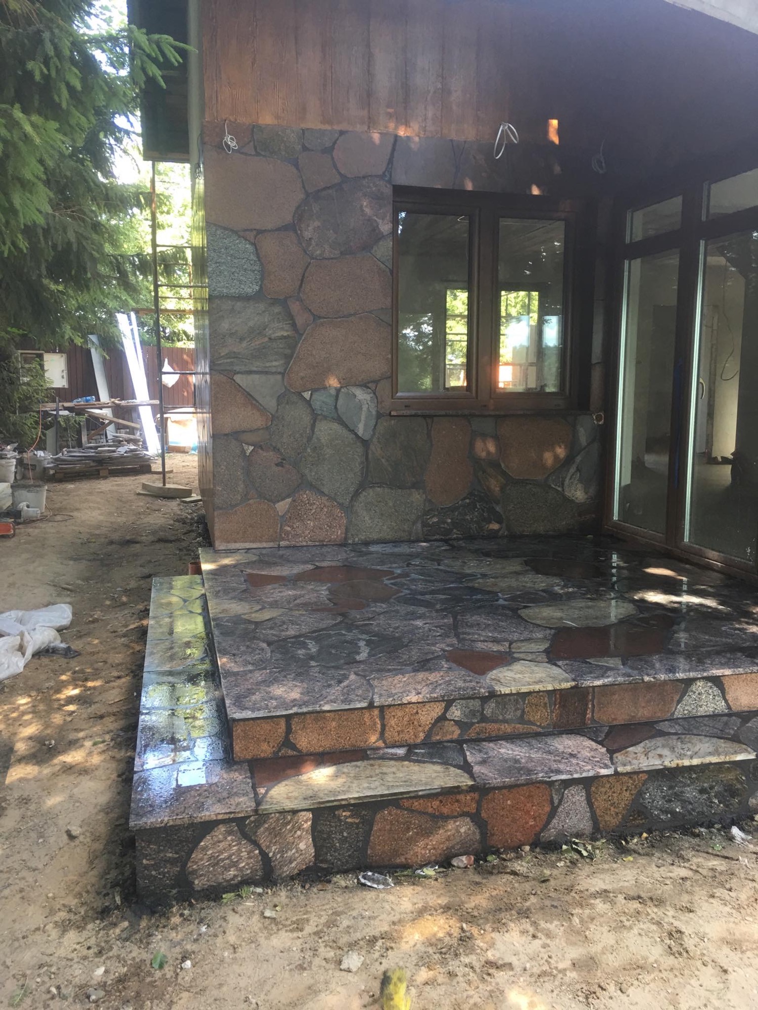 Kamienne schody wejściowe do domu, z widoczną elewacją z kamienia i drewnianym wykończeniem, w trakcie prac wykończeniowych na zewnątrz budynku.