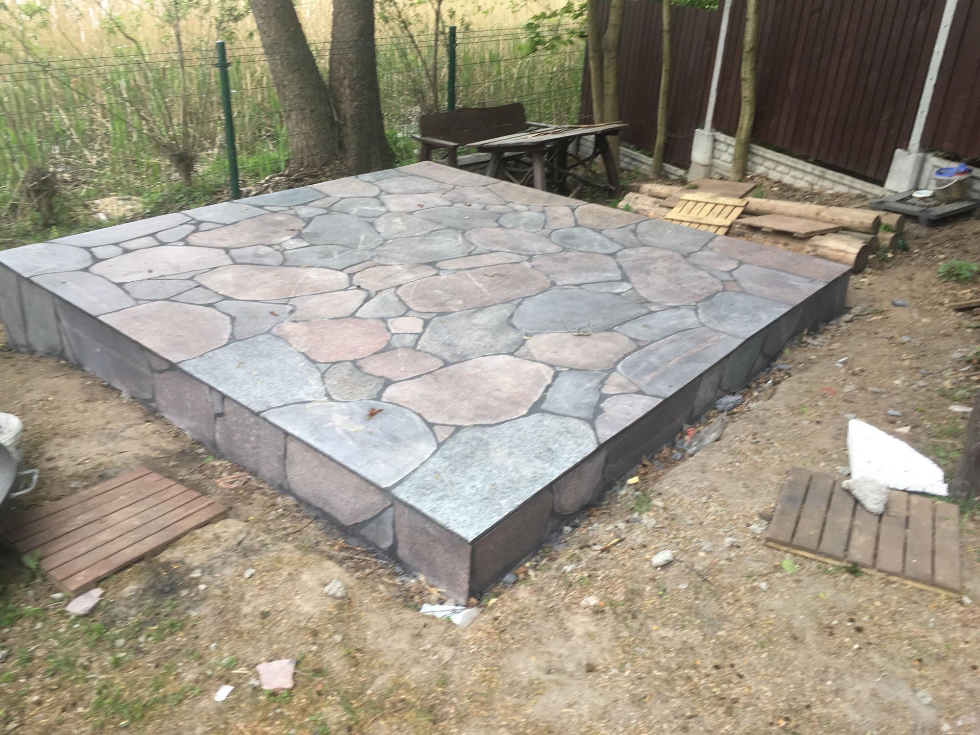 Kwadratowy taras z nieregularnych kamieni w odcieniach szarości i brązu, otoczony obrzeżem z szarej kostki, na tle ziemi i drzew.