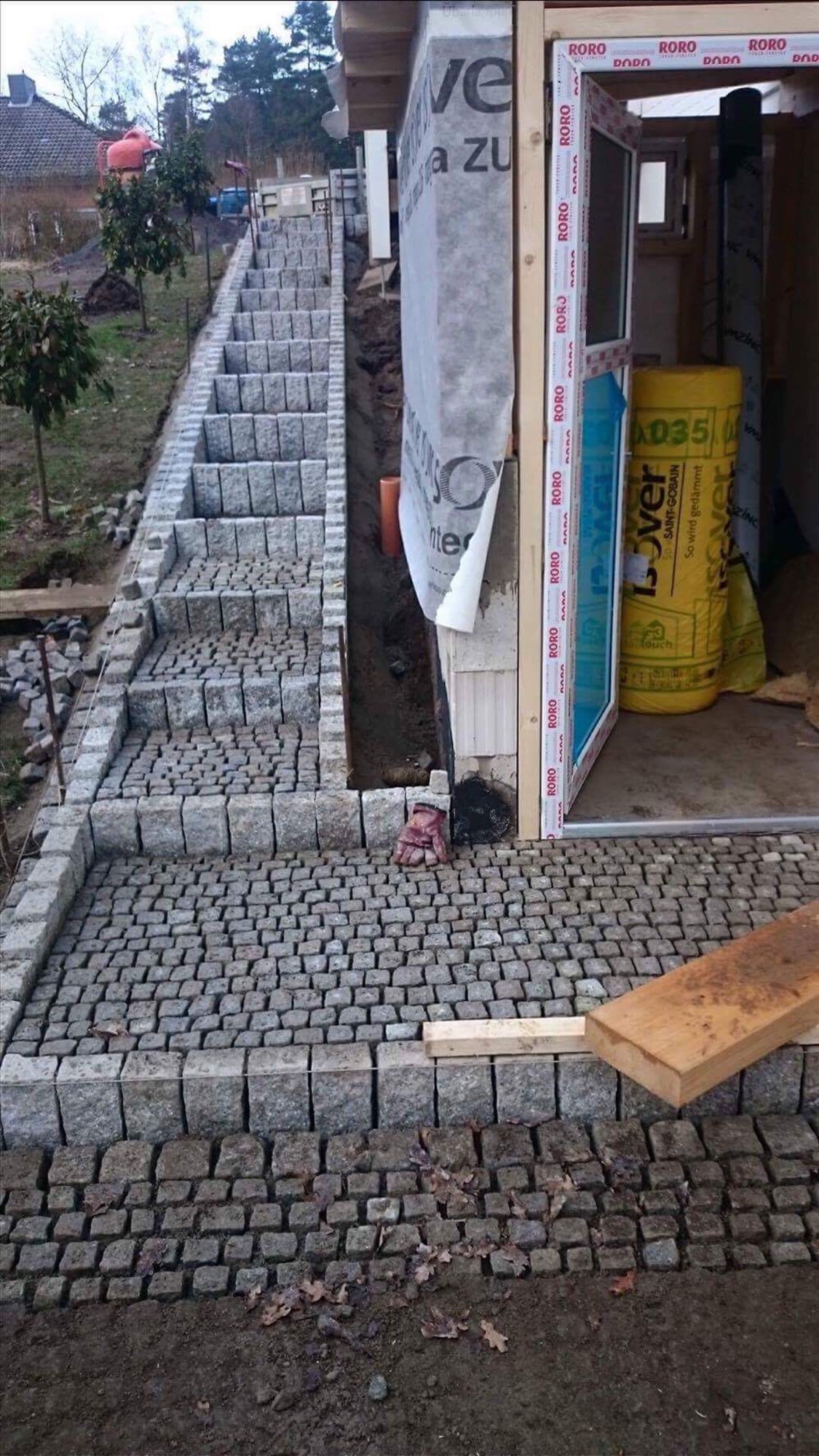 Schody zewnętrzne z kostki granitowej prowadzące w górę, częściowo ułożone, z widocznymi elementami konstrukcyjnymi budynku w tle i roboczą rękawicą obok wejścia.