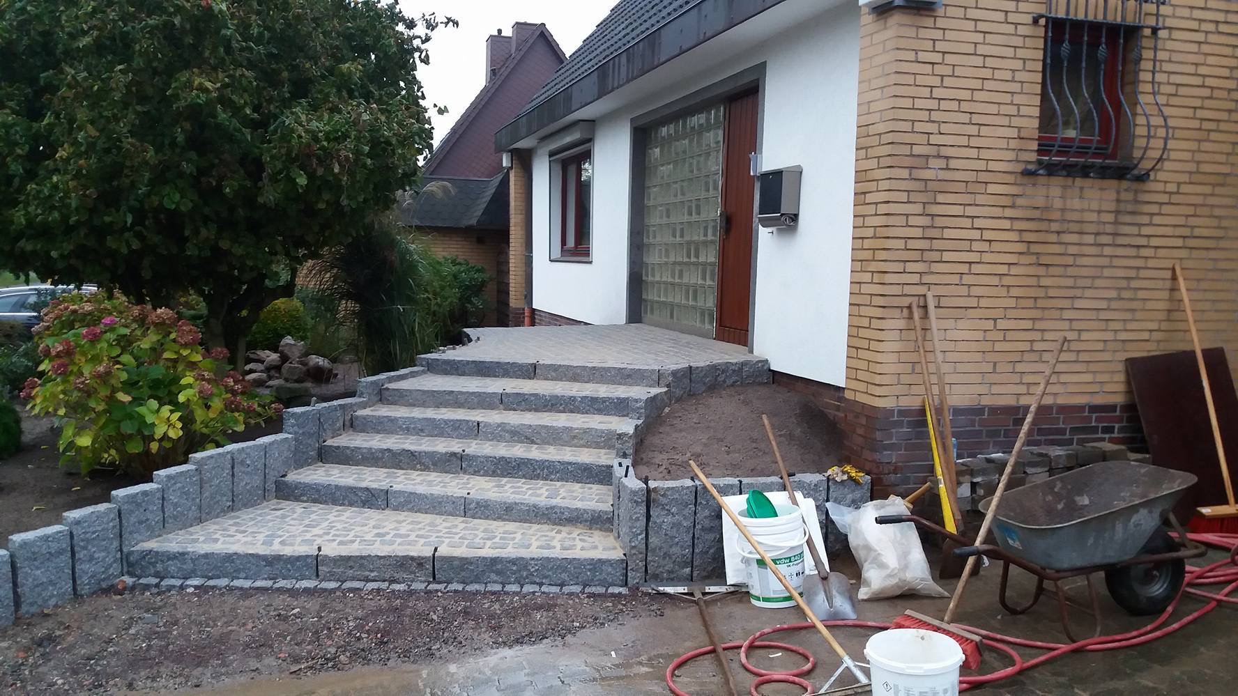 Granitowe schody prowadzące do domu z wejściem wyłożonym kostką, widoczne narzędzia budowlane i materiały wykończeniowe.