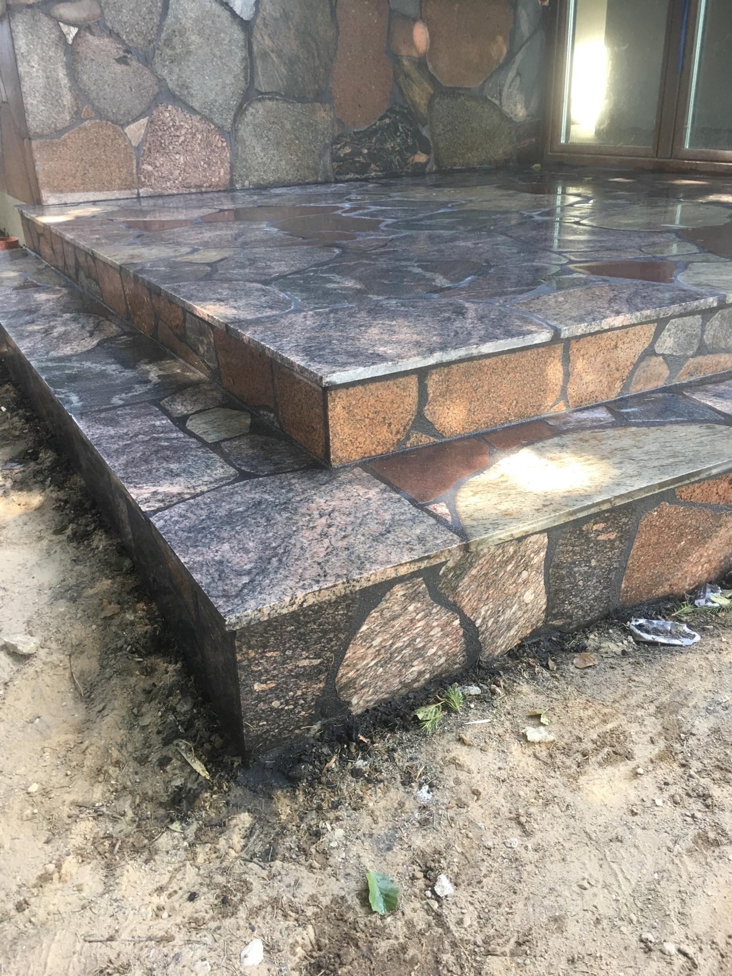 Dwu-stopniowe schody zewnętrzne wyłożone nieregularnymi płytami z polerowanego kamienia naturalnego w odcieniach szarości i brązu, widoczne mokre od świeżego mycia, z fragmentem elewacji budynku...