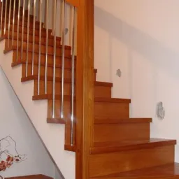 Schody drewniane z metalowymi balustradami, widoczne z dołu, z koszem wiklinowym i fragmentem butów u podstawy.