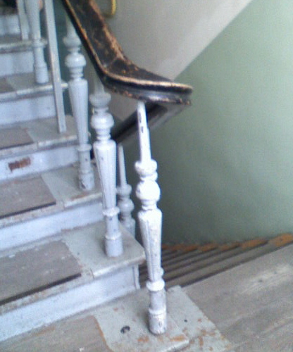 Schody drewniane z pomalowanymi na szaro stopniami i białą balustradą z ciemnobrązową poręczą, widoczne ślady użytkowania i odchodząca farba.