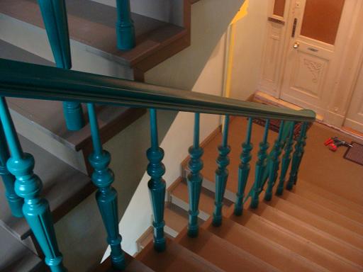 Drewniane schody z pomalowaną na turkusowo balustradą, widok z góry, w tle drzwi z dekoracyjnymi elementami i wkrętarka leżąca na podłodze.