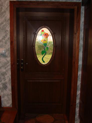 Ciemnobrązowe drzwi z owalnym, szklanym panelem przedstawiającym stylizowany kwiat z zielonym pędem, zamontowane w drewnianej futrynie.