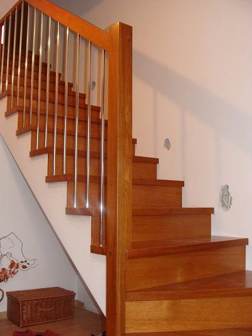 Schody drewniane z metalowymi balustradami, widoczne z dołu, z koszem wiklinowym i fragmentem butów u podstawy.