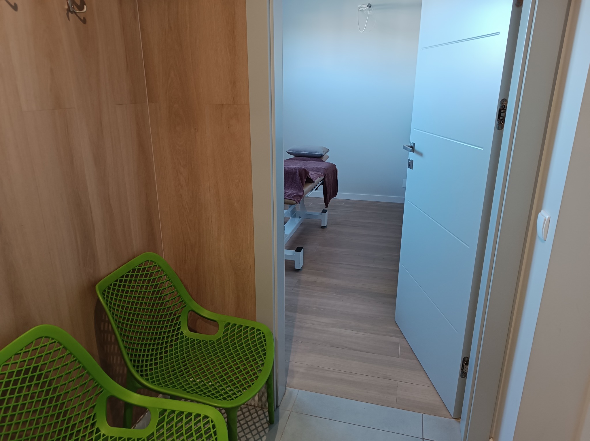 Jasne wnętrze gabinetu z zielonymi krzesłami, widok na łóżko do masażu przez otwarte drzwi. Minimalistyczny design, drewniane panele i subtelne oświetlenie.