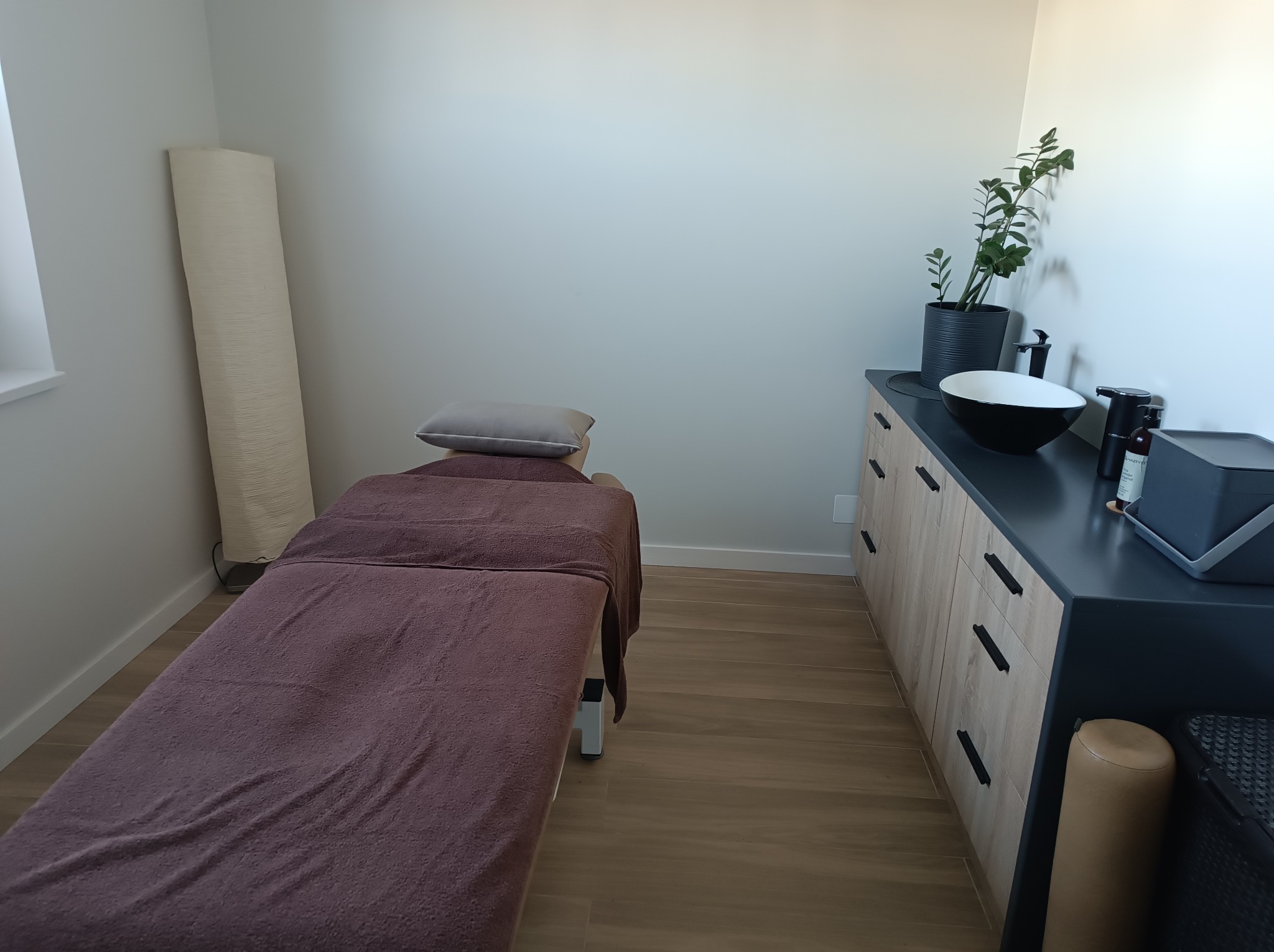 Jasne wnętrze gabinetu masażu z łóżkiem przykrytym brązowym ręcznikiem, umywalką i rośliną doniczkową. Minimalistyczny wystrój, drewniana podłoga.