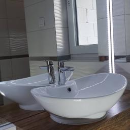 Firma ogólnobudowlana Grzegorz Żółtaszek - Dwie białe umywalki nablatowe o owalnym kształcie z chromowanymi bateriami, ustawione na blacie z drewnopodobnego materiału, widoczne na tle ściany z jasnych płytek z delikatnym wzorem i lustra...