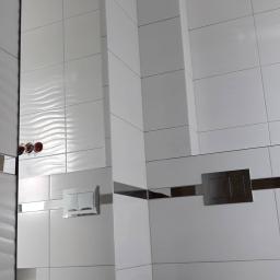 Firma ogólnobudowlana Grzegorz Żółtaszek - Nowoczesna łazienka wykończona białymi płytkami z dekoracyjnym wzorem fal i akcentami w kolorze chromu oraz ciemnego drewna, widoczny przycisk spłuczki.