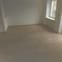 Jasny parkiet ułożony w pustym pokoju z białymi ścianami, widoczne okno i drzwi balkonowe z widokiem na ogród.