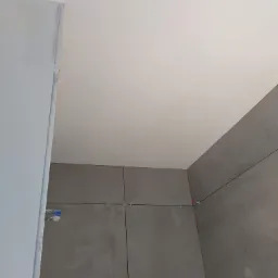 Szare płytki wielkoformatowe układane na ścianie w rogu pomieszczenia, widoczne krzyżyki dystansowe i fragment sufitu oraz pomalowana ściana z listwą wykończeniową.