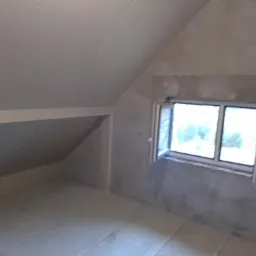 Pokój na poddaszu w trakcie prac wykończeniowych, widoczne surowe ściany, okno z otwartym skrzydłem i przygotowane podłoże pod podłogę.