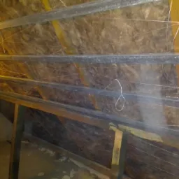 Poddasze w trakcie ocieplania wełną mineralną, widoczne metalowe profile i przymocowana sznurkiem folia paroizolacyjna, oświetlenie robocze.