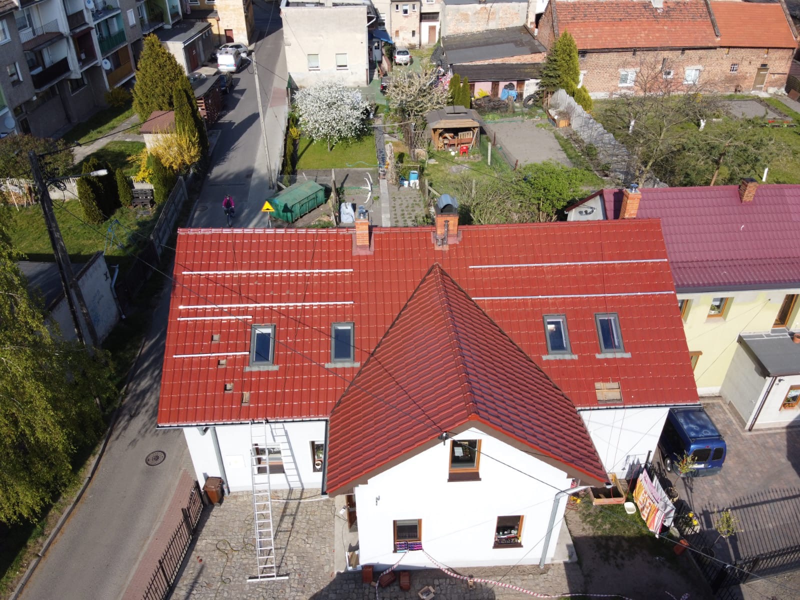 Czerwony dach z zamontowanymi profilami pod panele słoneczne widziany z góry; biała fasada budynku z oknami dachowymi, drabina oparta o ścianę.