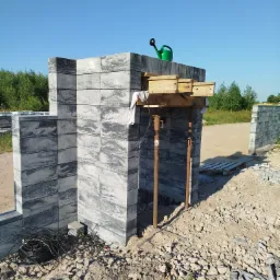 Wznoszenie muru z szarych bloczków betonowych z widocznymi podporami drewnianymi i metalowymi, na górze muru stoi zielona konewka. W tle widoczny teren budowy i zieleń.