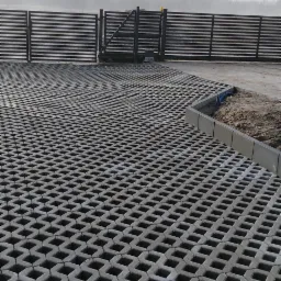 Ułożona szara ażurowa nawierzchnia z betonowych płyt, brama wjazdowa i poziome przęsła ogrodzenia w tle, pochmurny dzień.