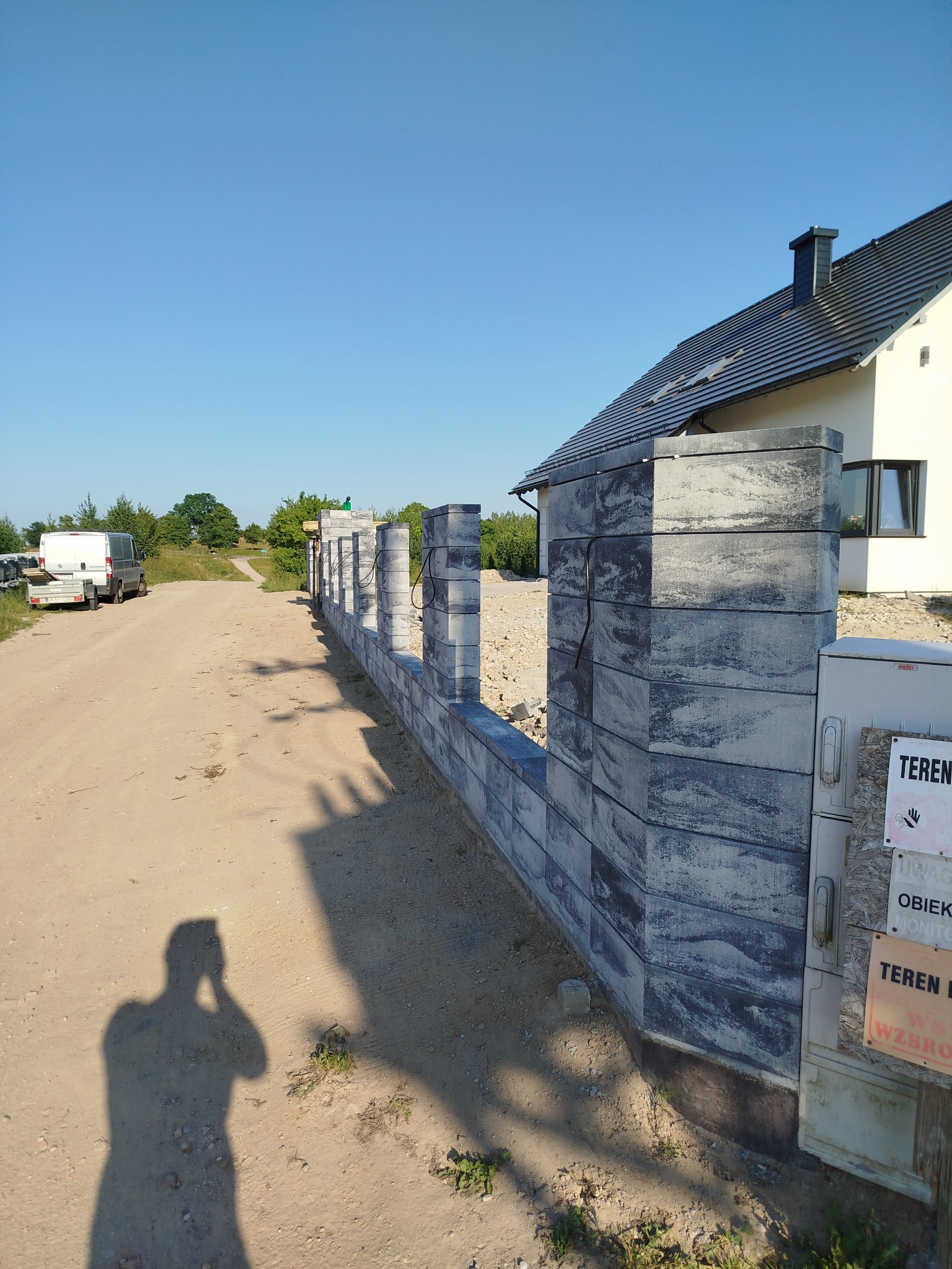 Wznoszone ogrodzenie z szarych, betonowych bloczków w trakcie budowy, widoczny cień fotografa, dom w tle, teren budowy.