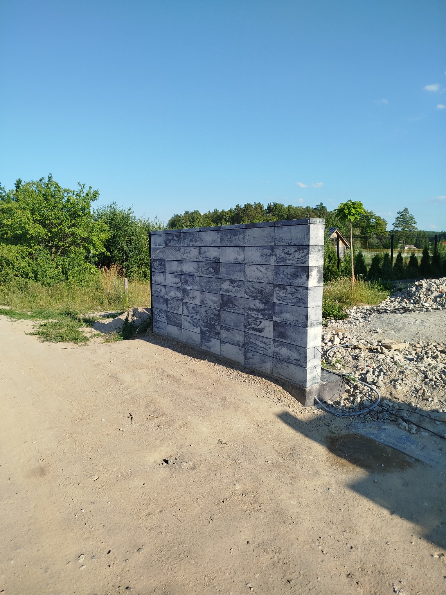 Fragment muru z szarej kostki betonowej, prawdopodobnie element ogrodzenia, na piaszczystym podłożu z widocznymi kablami, w tle zieleń i błękitne niebo.