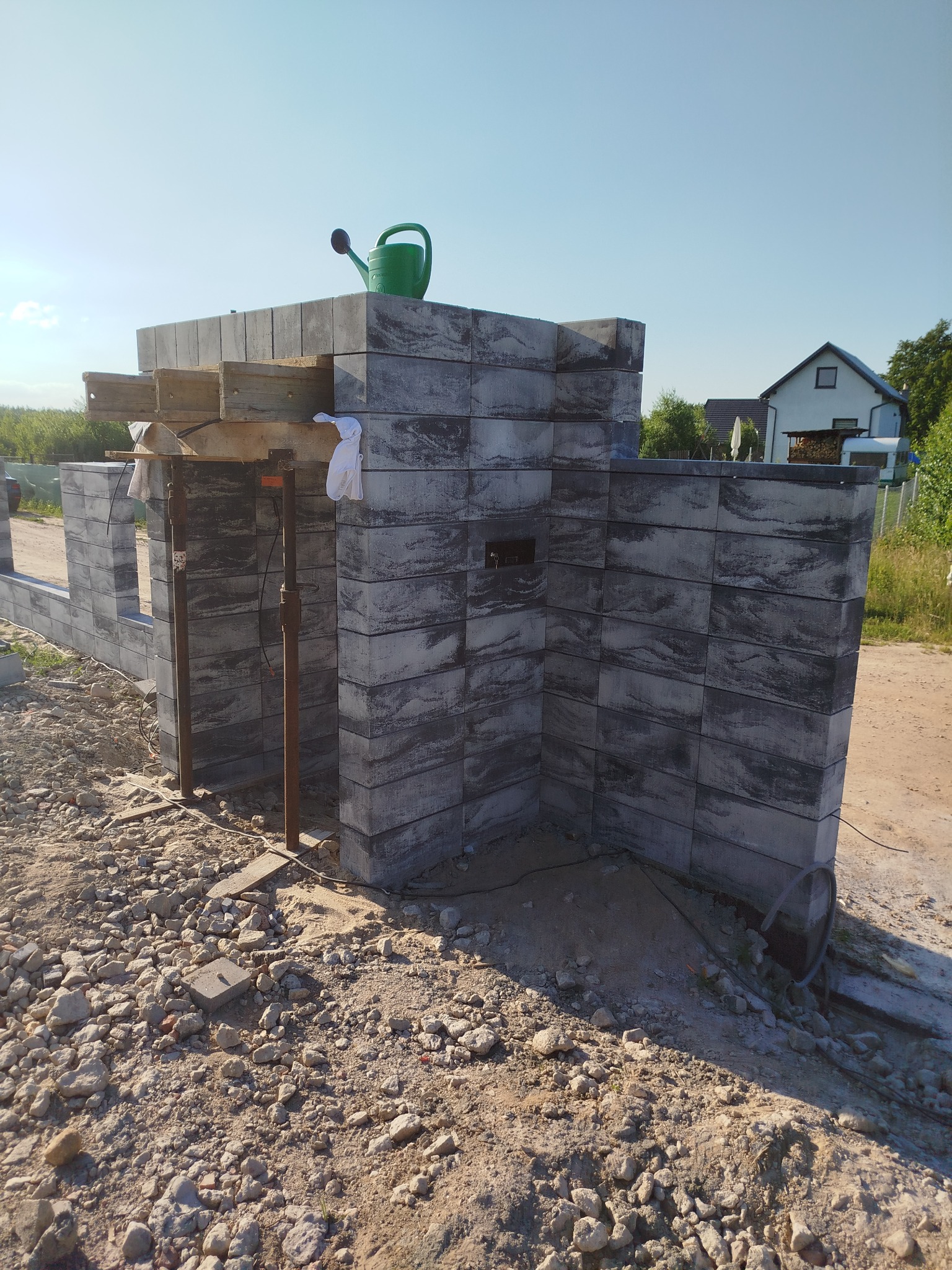 Wznoszona konstrukcja z szarych, betonowych bloczków z widoczną fakturą, podparta drewnianymi belkami i metalowymi stemplami; na górze stoi zielona konewka.