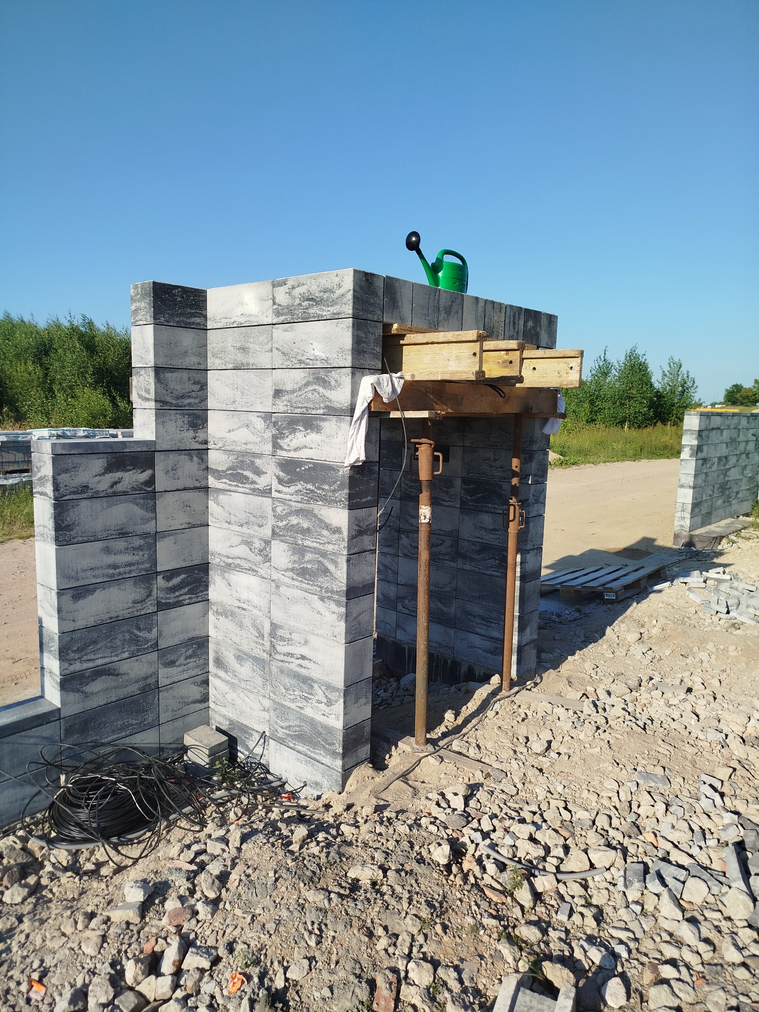 Wznoszenie muru z szarych bloczków betonowych z widocznymi podporami drewnianymi i metalowymi, na górze muru stoi zielona konewka. W tle widoczny teren budowy i zieleń.