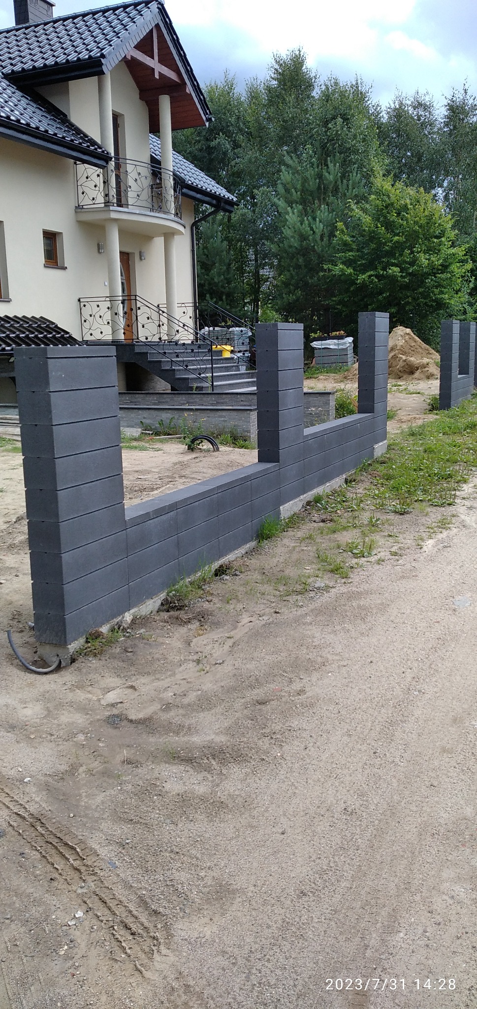 Wznoszenie szarego ogrodzenia z betonowych bloczków na posesji, widoczny fragment domu z balkonem w tle, etap budowy.