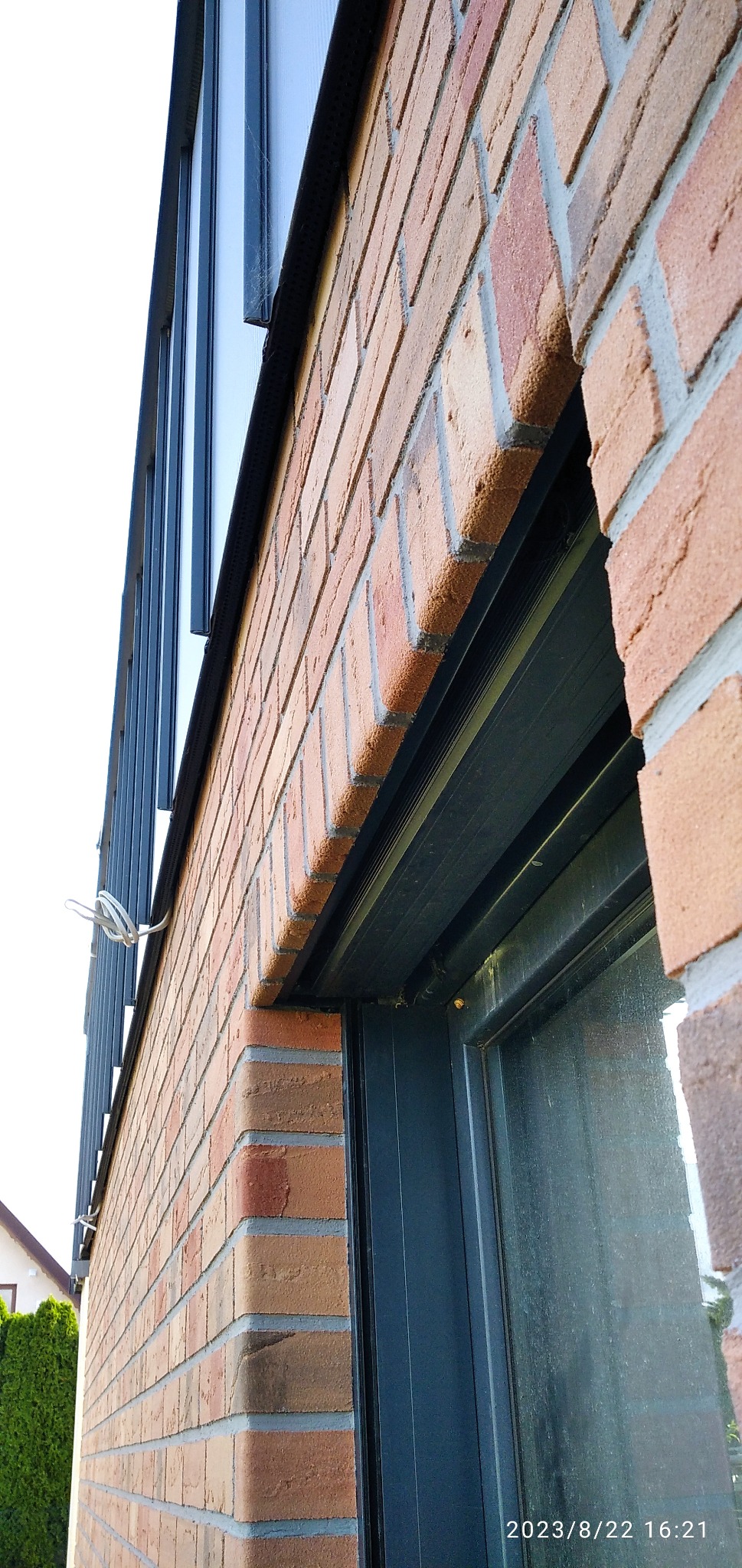 Fragment murowanej ściany budynku z czerwonej cegły, widoczne okno z ciemną ramą oraz fragment okna dachowego z bliska, z zewnątrz budynku.