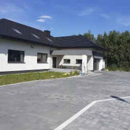 Nowoczesny dom jednorodzinny z szarą kostką brukową na podjeździe, białą elewacją i czarną dachówką, widoczne narzędzia i drabina sugerujące prace wykończeniowe.