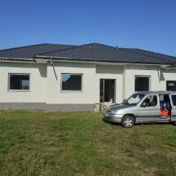 Nowo wybudowany dom jednorodzinny z szarą dachówką i świeżo pomalowaną białą elewacją, widoczny fragment ozdobnej podmurówki, na trawniku zaparkowany srebrny samochód z otwartymi drzwiami.